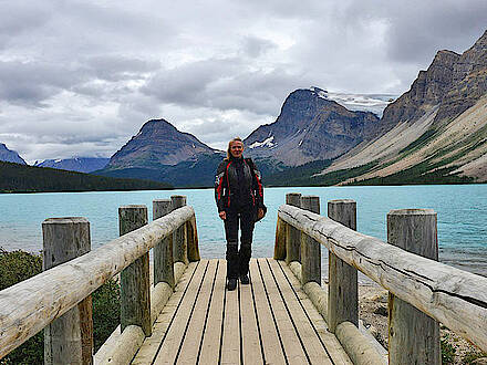 Motorradfahrerin steht auf einem Steg an einem See am Icefield Parkwal mit Bergen im Hintergrund in Kanada